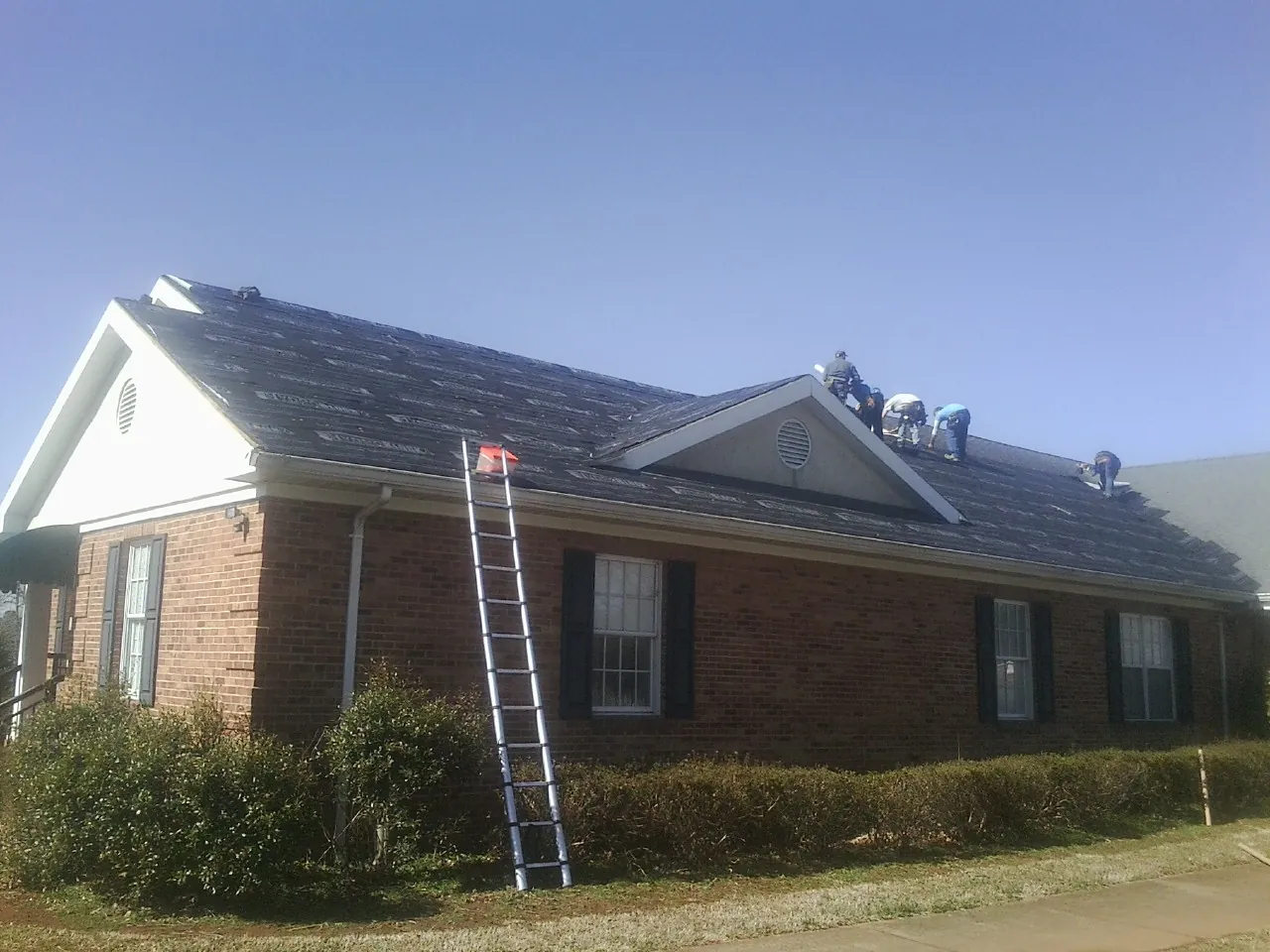 Crew tearing shingles off a brick ranch roof with a ladder set — Emergency Roof Repair work by our crew in Hewlett