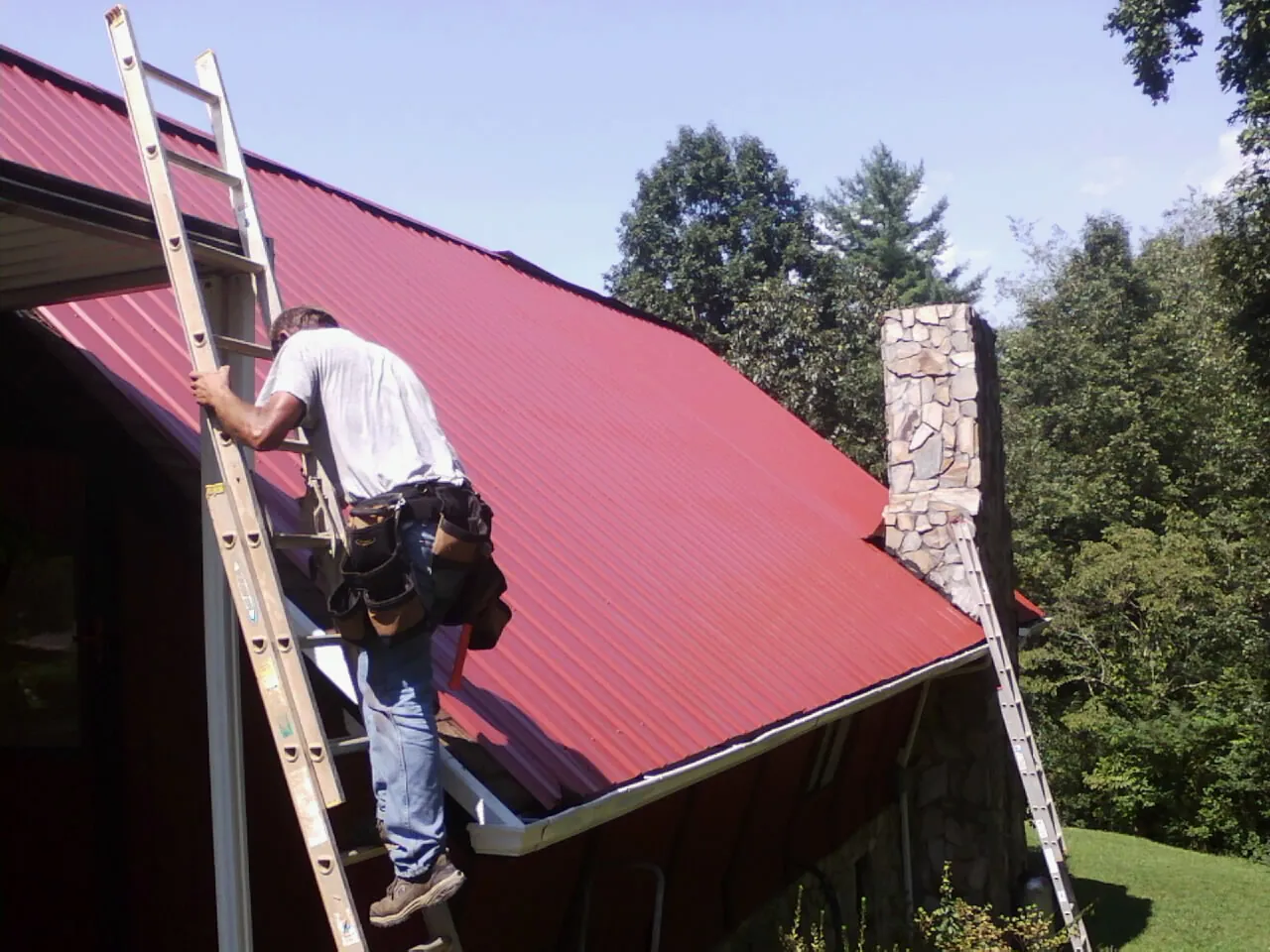 Roofer on a ladder beside a red metal roof and stone chimney — Soffit & Fascia Repair by Roofing Brothers in Hewlett